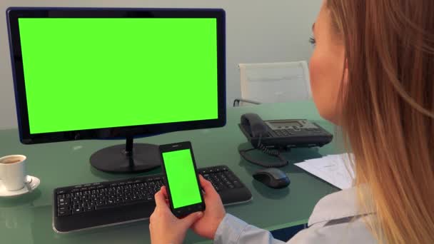 Uma mulher senta-se em um escritório e olha em turnos para uma tela de computador e um smartphone (ambos tela verde ) — Vídeo de Stock