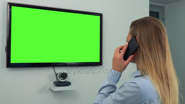Una mujer habla en un teléfono inteligente frente a una pantalla de televisión verde — Vídeo de stock