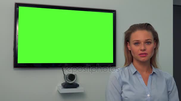 Молодая, красивая женщина смотрит в камеру, зеленый телевизор на заднем плане — стоковое видео