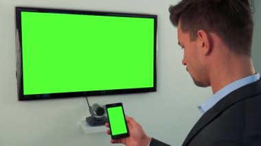 Bakan bir adam sırayla yeşil televizyon ekran ve bir akıllı telefon ile yeşil bir ekran elinde
