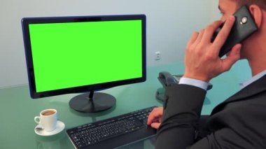 Bir adam bir yeşil ekran ve bir ofiste bir akıllı telefon üzerinde konuşmak olan bir bilgisayarda çalışır