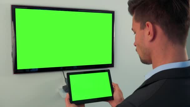 Un hombre se para frente a una pantalla de televisión verde y mira a su vez a la televisión y una tableta con una pantalla verde — Vídeo de stock