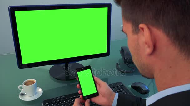 Человек сидит в офисе и смотрит в очереди на экран компьютера и смартфон (и зеленый экран ) — стоковое видео