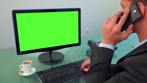 Un hombre trabaja en una computadora con una pantalla verde y habla en un teléfono inteligente en una oficina — Vídeo de stock