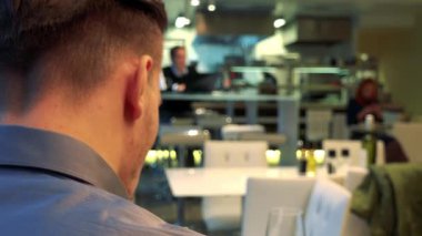 Bir adam (başının arkasına kameraya) bir restoranda bir masada oturuyor, yiyor ve Şarap içer