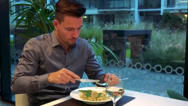 Молодой, красивый мужчина сидит в ресторане и ест макароны, бокал вина перед ним — стоковое видео
