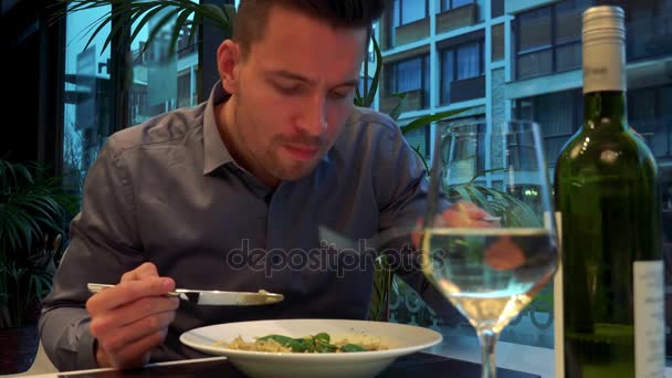 若く、ハンサムな男がレストランでテーブルに座っているし、フォア グラウンドでパスタ、ボトル、ワインのガラスを食べる — ストック動画