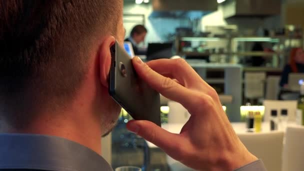 Een man (de achterkant van zijn hoofd naar de camera) zit aan een tafel in een restaurant en praat op een smartphone - closeup — Stockvideo