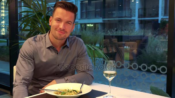 Un hombre joven y guapo se sienta en una mesa en un restaurante y sonríe a la cámara, su comida y su copa de vino en la mesa. — Vídeos de Stock