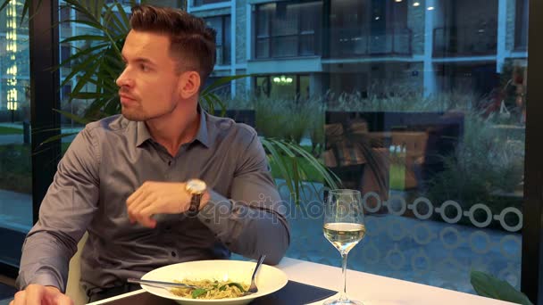 Egy fiatal, jóképű férfi ül egy asztalnál, egy étteremben, és türelmetlenül vár — Stock videók