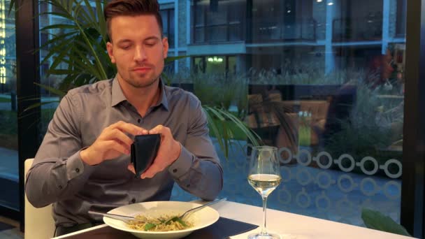 Ein junger, gutaussehender Mann sitzt an einem Tisch in einem Restaurant, hält ein Portemonnaie und zählt — Stockvideo