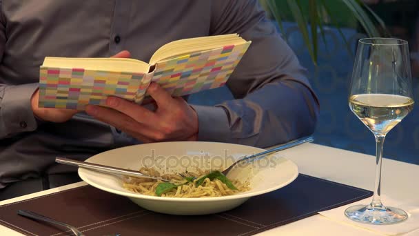 Egy férfi ül egy asztalnál, egy étteremben, és olvas egy könyvet, ételt és pohár bort az asztalon — Stock videók