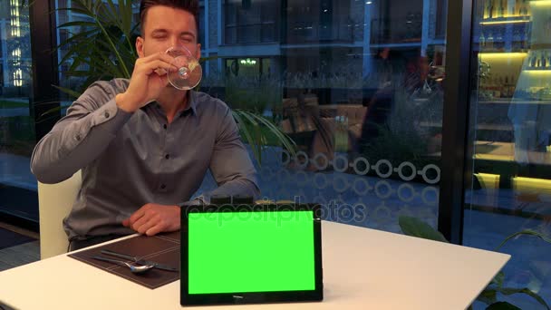 Un uomo giovane e bello si siede a un tavolo in un ristorante, beve vino e sorride alla macchina fotografica - un tablet con uno schermo verde in primo piano sul tavolo — Video Stock