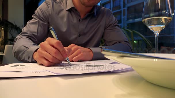Um homem senta-se a uma mesa em um restaurante e escreve no papel, um prato em primeiro plano — Vídeo de Stock