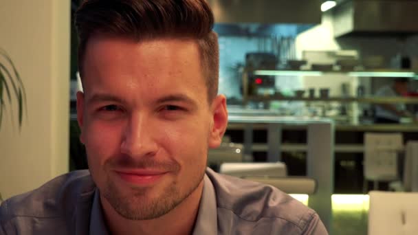 Un hombre joven y guapo se sienta en una mesa en un restaurante y sonríe a la cámara - de cerca — Vídeos de Stock
