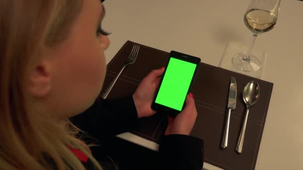Een vrouw zit aan een tafel in een restaurant en kijkt naar een smartphone met een groen scherm — Stockvideo