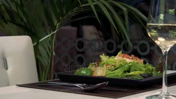 Una cena sabrosa (ensalada con nueces y una copa de vino blanco) en una mesa en un restaurante — Vídeos de Stock