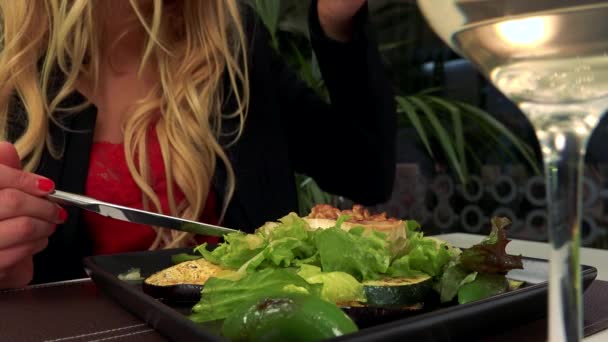 Una mujer se sienta en una mesa en un restaurante y come una ensalada, una copa de vino en primer plano — Vídeo de stock