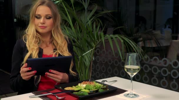 Uma jovem e bela mulher senta-se a uma mesa em um restaurante e trabalha em um tablet, uma salada e um copo de vinho na frente dela — Vídeo de Stock