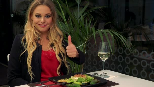 Uma jovem e bela mulher senta-se à mesa de um restaurante e mostra um polegar até a câmera, uma refeição na frente dela — Vídeo de Stock