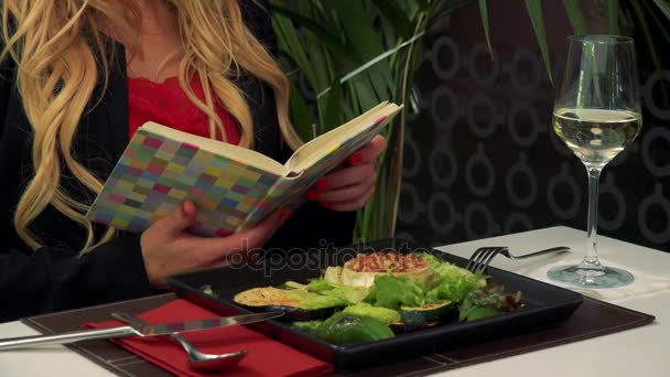 Μια γυναίκα που κάθεται σε ένα τραπέζι σε ένα εστιατόριο και διαβάζει ένα βιβλίο, μια σαλάτα μπροστά της — Αρχείο Βίντεο