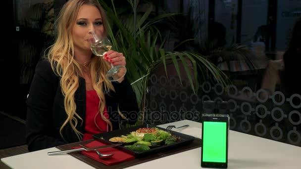 Une jeune et belle femme s'assoit à une table dans un restaurant, sourit à la caméra et boit du vin - un smartphone avec un écran vert sur la table — Video