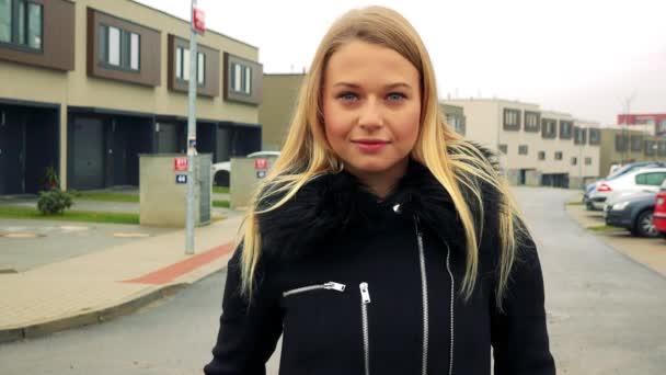 Una giovane, bella donna si trova sulla strada e guarda la macchina fotografica, edifici sullo sfondo — Video Stock