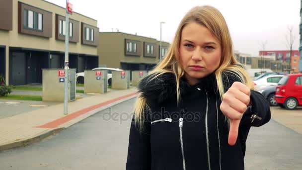 A young, beautiful woman stands on the street in a suburban area, shows a thumb down to the camera and shakes her head — Stock Video
