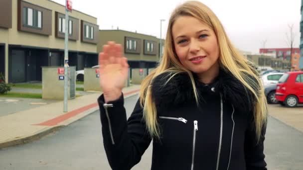 Uma jovem, bela mulher fica na rua em uma área suburbana, sorrisos e ondas mão para a câmera — Vídeo de Stock