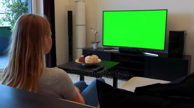 Sarışın bir kadın rahat bir oturma odasında bir kanepede oturuyor ve yeşil bir ekran bir Tv izliyor