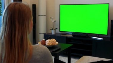 Sarışın kadın (başının arkasına kameraya) rahat bir oturma odasında bir kanepede oturuyor ve yeşil bir ekran bir Tv izliyor