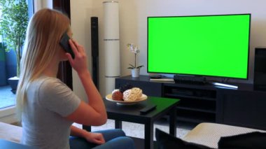 Sarışın bir kadın bir kanepe oturma odasında oturur ve smartphone cep telefonu ile arka planda yeşil bir ekran bir Tv görüşmeler