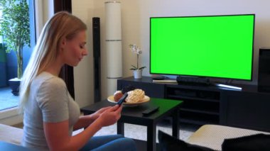 Genç, güzel bir kadın bir Tv ile bir oturma odası ve çalışan bir smartphone üzerinde yeşil bir ekran önünde bir kanepede oturuyor