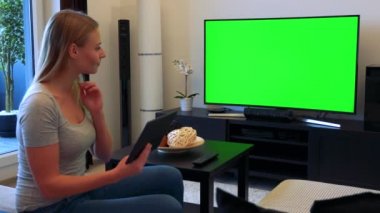 Genç, güzel bir kadın bir Tv ile bir oturma odası ve bir tablet üzerinde çalışan yeşil bir ekran önünde bir kanepede oturuyor