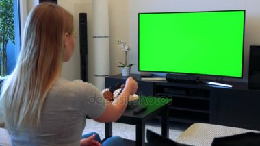 Sarışın bir kadın bir oturma odasında bir kanepede oturuyor, yeşil bir ekran bir Tv izliyor ve bir Tv denetleyicisini kullanır