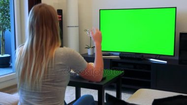 Sarışın bir kadın bir Tv ile bir oturma odasında yeşil bir ekran önünde bir kanepede oturuyor, alkış ve sonra kutluyor