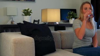 Genç, güzel bir kadın bir oturma odasında bir kanepede oturuyor, bir bardak su içer ve kamera kapalı olan bir Tv izliyor