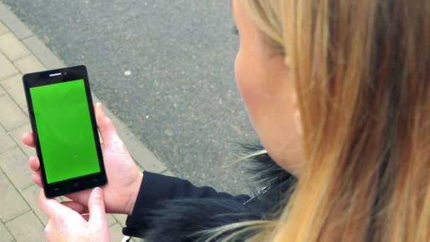 Een blonde vrouw staat op een stoep en kijkt naar een smartphone met een groen scherm — Stockvideo