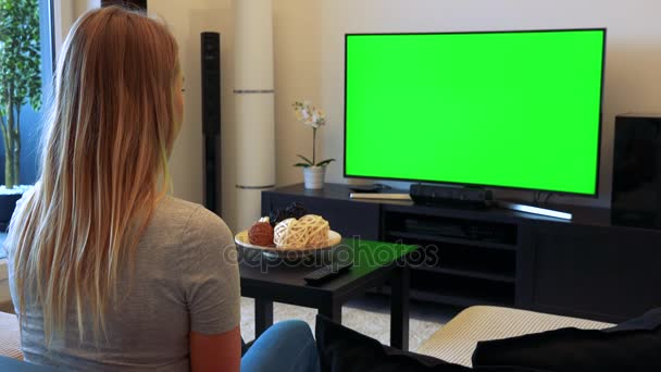 Uma jovem, bela mulher senta-se em um sofá em uma sala de estar e assiste a uma TV com uma tela verde, em seguida, vira e sorri para a câmera — Vídeo de Stock