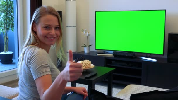 Uma jovem, bela mulher senta-se em um sofá em uma sala de estar e assiste a uma TV com uma tela verde, em seguida, vira e mostra um polegar para a câmera com um sorriso — Vídeo de Stock