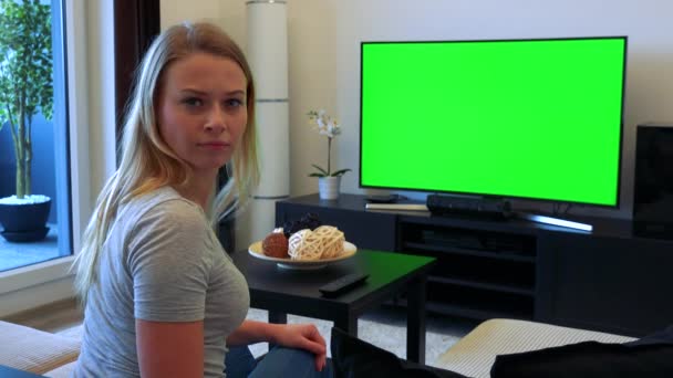 Eine junge, schöne Frau sitzt auf einer Couch im Wohnzimmer und sieht einen Fernseher mit grünem Bildschirm, wendet sich dann der Kamera zu und schüttelt den Kopf — Stockvideo