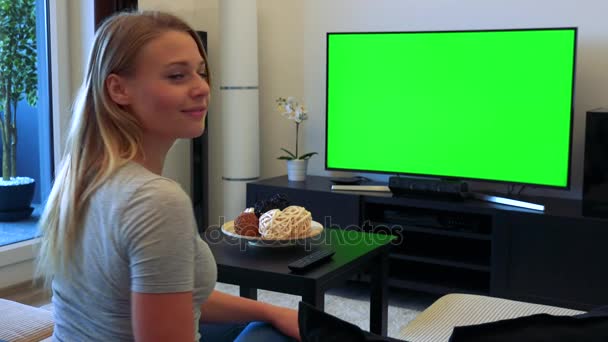 Uma jovem e bela mulher senta-se em um sofá em uma sala de estar e assiste a uma TV com uma tela verde, depois se vira para a câmera e acena com um sorriso — Vídeo de Stock