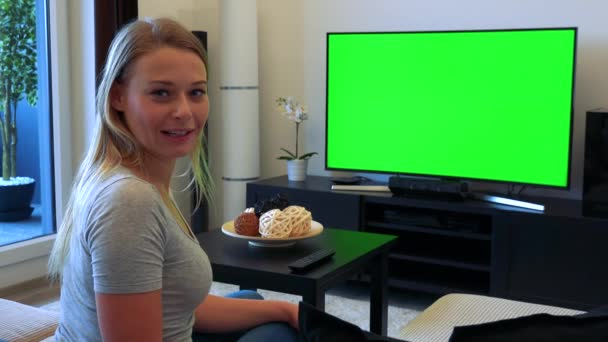 Una giovane, bella donna si siede su un divano in un soggiorno e guarda una TV con uno schermo verde, poi si gira e parla con la fotocamera — Video Stock