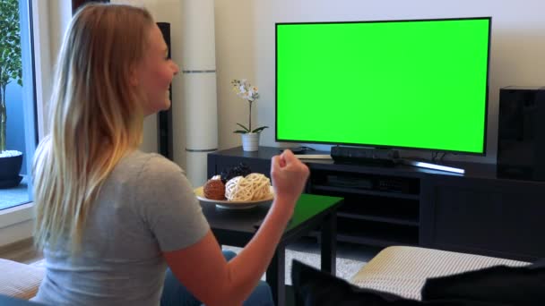 Eine blonde Frau sitzt auf einer Couch im Wohnzimmer, schaut einen Fernseher mit grünem Bildschirm und feiert — Stockvideo