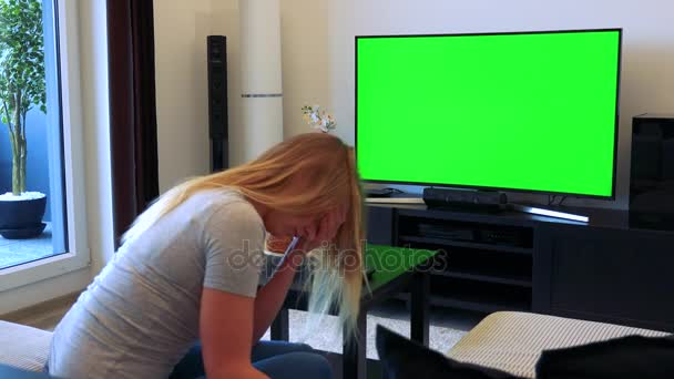 A blonde woman sits on a couch in a living room and watches a TV with a green screen, then covers her face with her hands unhappily — Stock Video