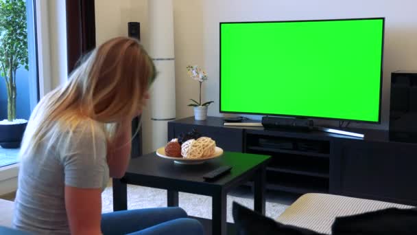 A blonde woman sits on a couch in a living room, watches TV with a green screen and gets angry — Stock Video