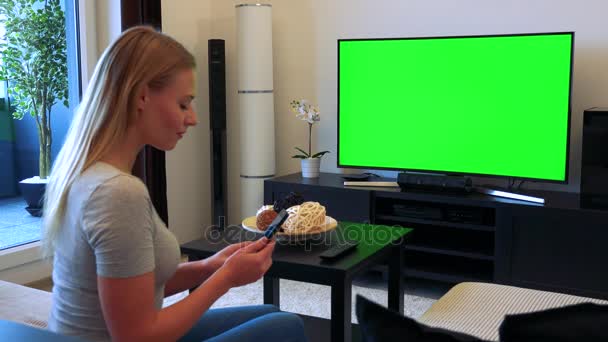 A young, beautiful woman sits on a couch in front of a TV with a green screen in a living room and works on a smartphone — Stock Video
