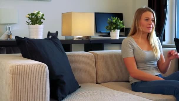 A young, beautiful woman sits on a couch in a living room, watches a TV which is off the camera and gets angry — Stock Video