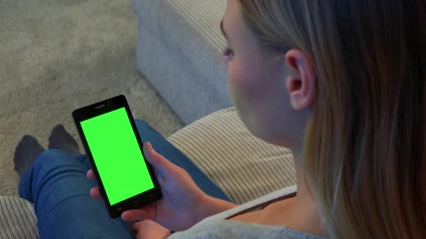 Blond žena sedí na gauči a v její ruce se dívá na smartphone s zeleným plátnem