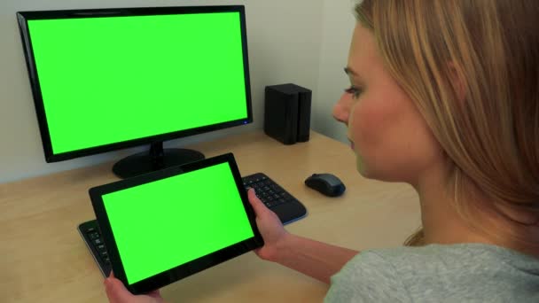 Uma jovem mulher senta-se em uma mesa e olha em turnos para uma tela de computador e um tablet (ambos tela verde ) — Vídeo de Stock